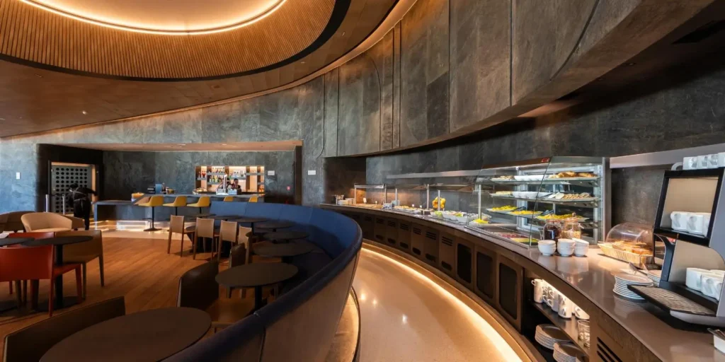 Sala VIP de aeroporto com buffet gastronômico, balcão de bebidas, mesas, poltronas confortáveis e ambiente sofisticado.
